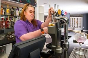 A barista at a coffee machine with multiple beer taps, a computer monitor, and a glass of iced drink on the counter. - Ibis Styles Haydock (St Helens)