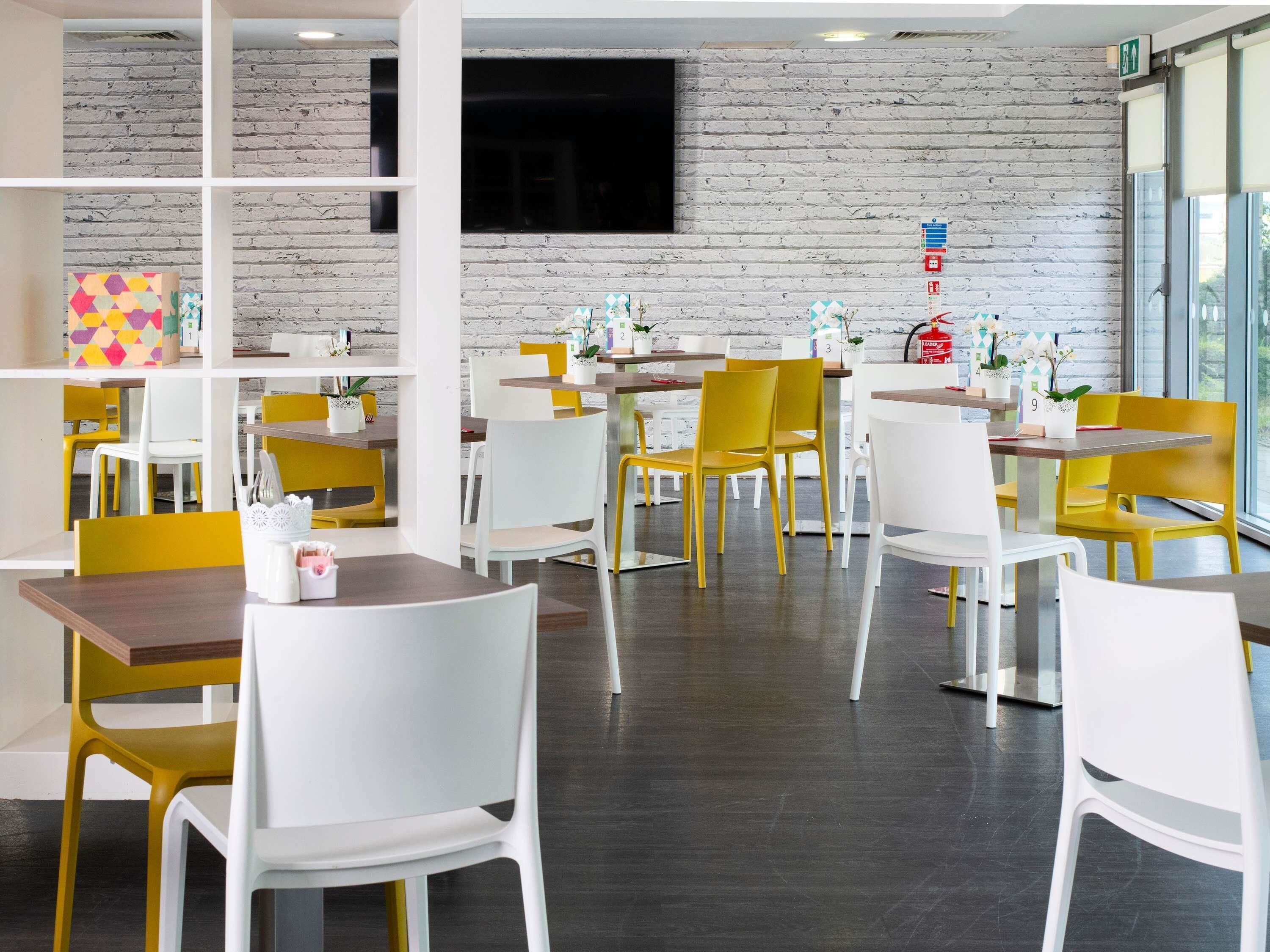 A modern dining area with white and yellow chairs, wooden tables, and a brick wall.