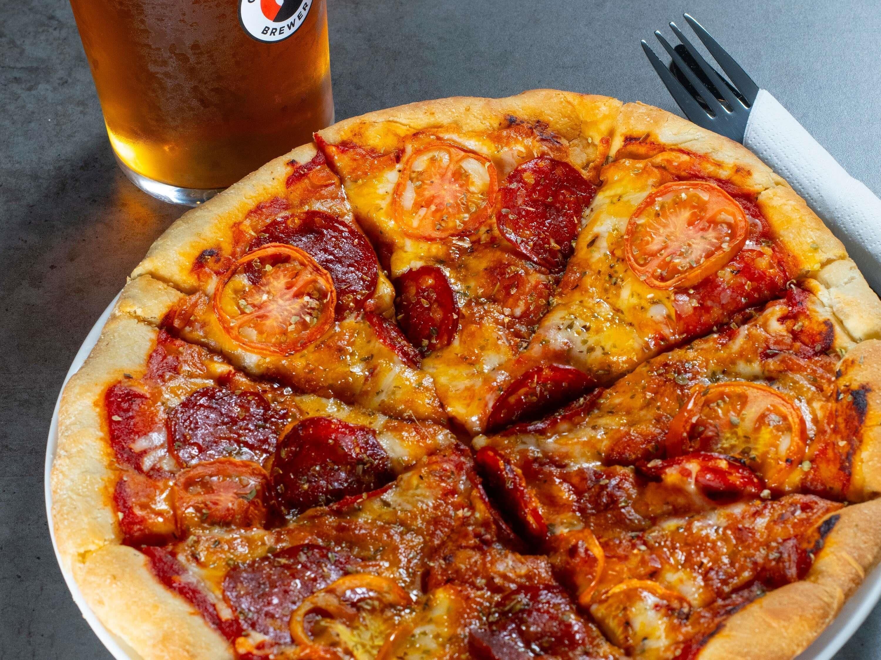 A pizza with tomato and pepperoni toppings, a beer glass, and a fork on a table.