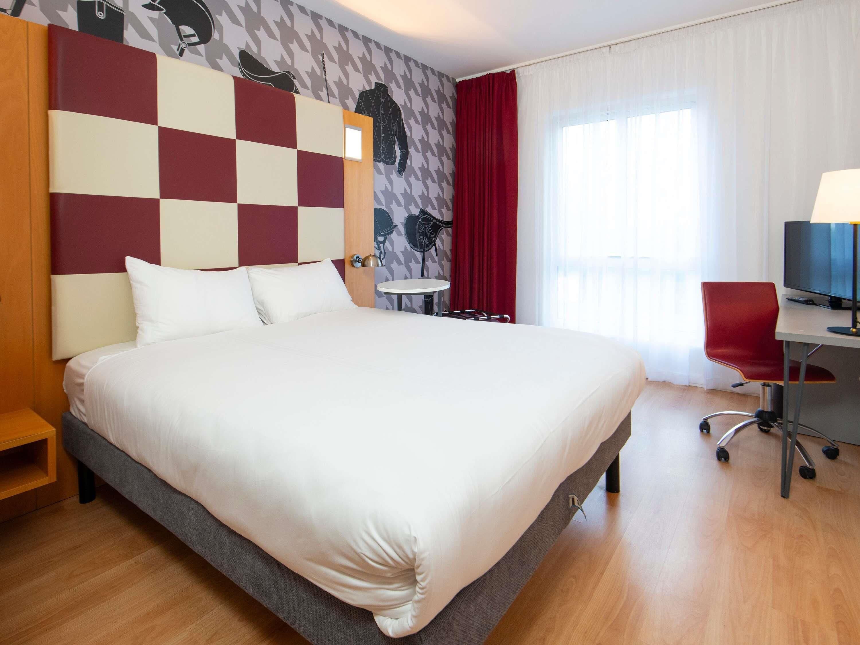 A hotel room with a large bed, a desk with a chair, a small table, and a TV.