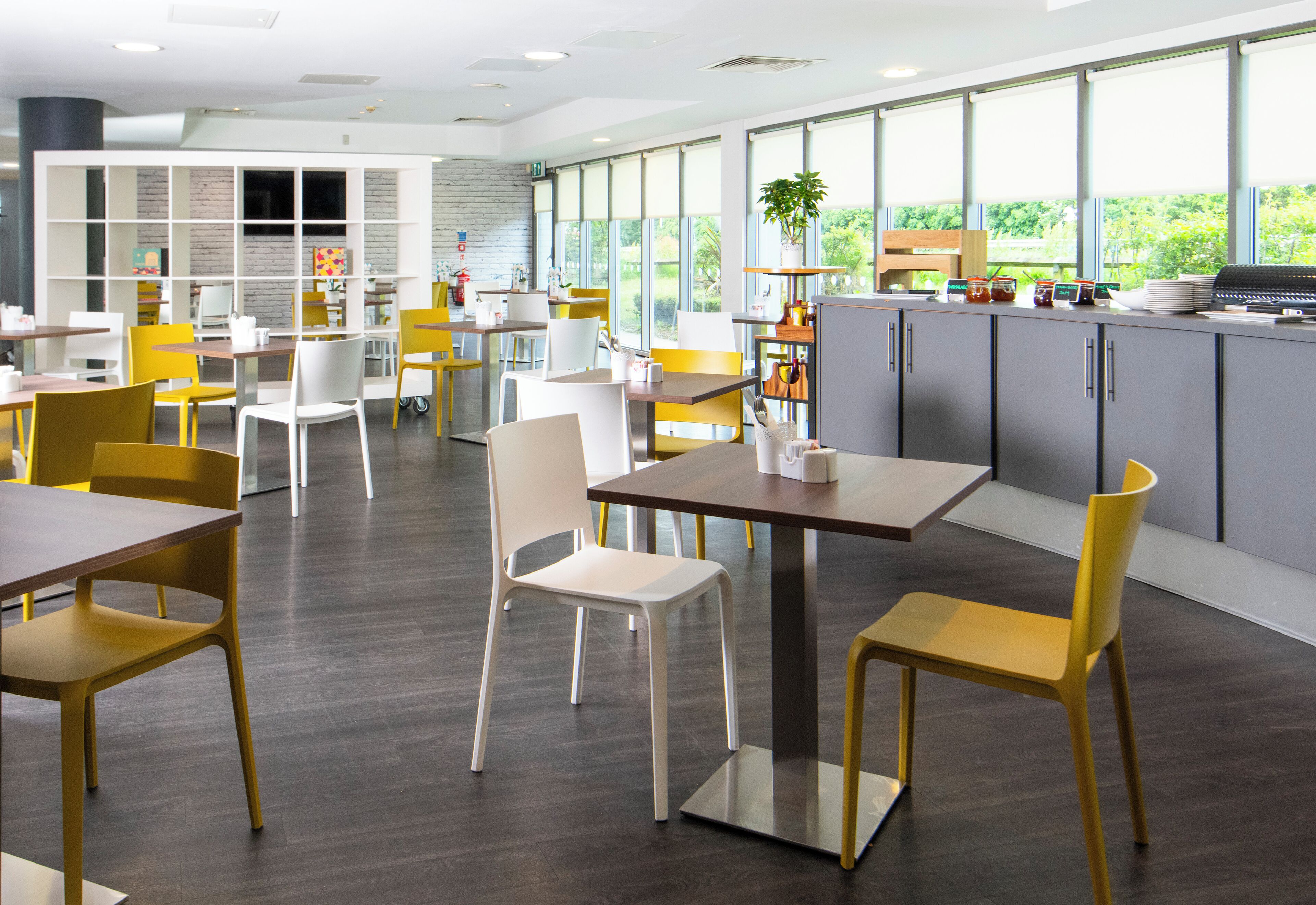 Uma área de refeições moderna com várias mesas e cadeiras, bancada de cozinha e grandes janelas com vista para o verde.
