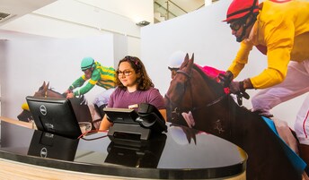 Una mujer frente a un escritorio con una computadora portátil y un monitor, en una habitación con un cartel grande de jinetes a caballo.