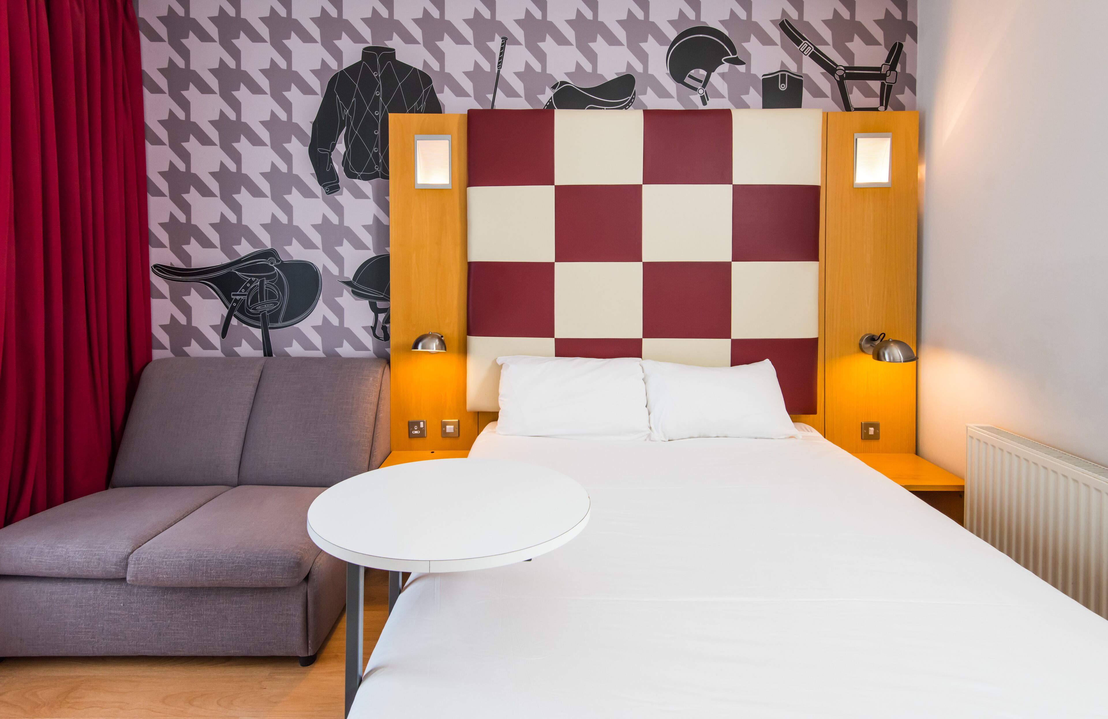 A hotel room with a checkered headboard, a grey sofa, a round white table, and a red curtain.