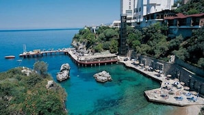 A coastal hotel with a pier extending into the sea, surrounded by lush greenery and clear blue waters. - Korumar Deluxe Hotel (Kusadasi)