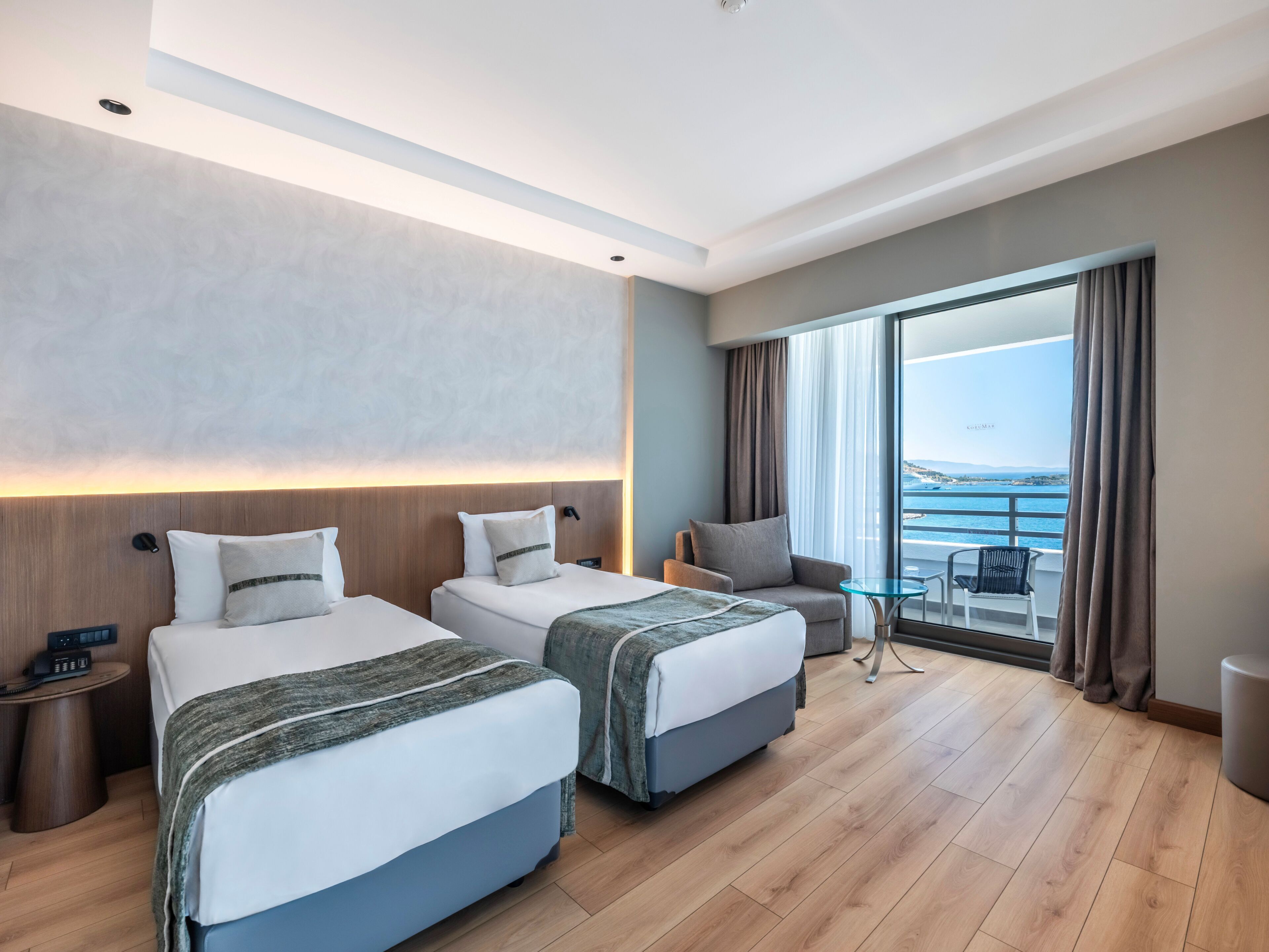 Ein Hotelzimmer mit zwei Betten, einem kleinen Tisch, einem Sessel und einem großen Fenster mit Meerblick.