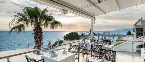 A terrace with tables set for dining, overlooking a body of water and mountains.