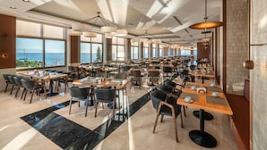 A spacious restaurant with multiple tables and chairs, large windows offering a view of the sea, and modern pendant lighting. - Korumar Deluxe Hotel (Kusadasi)