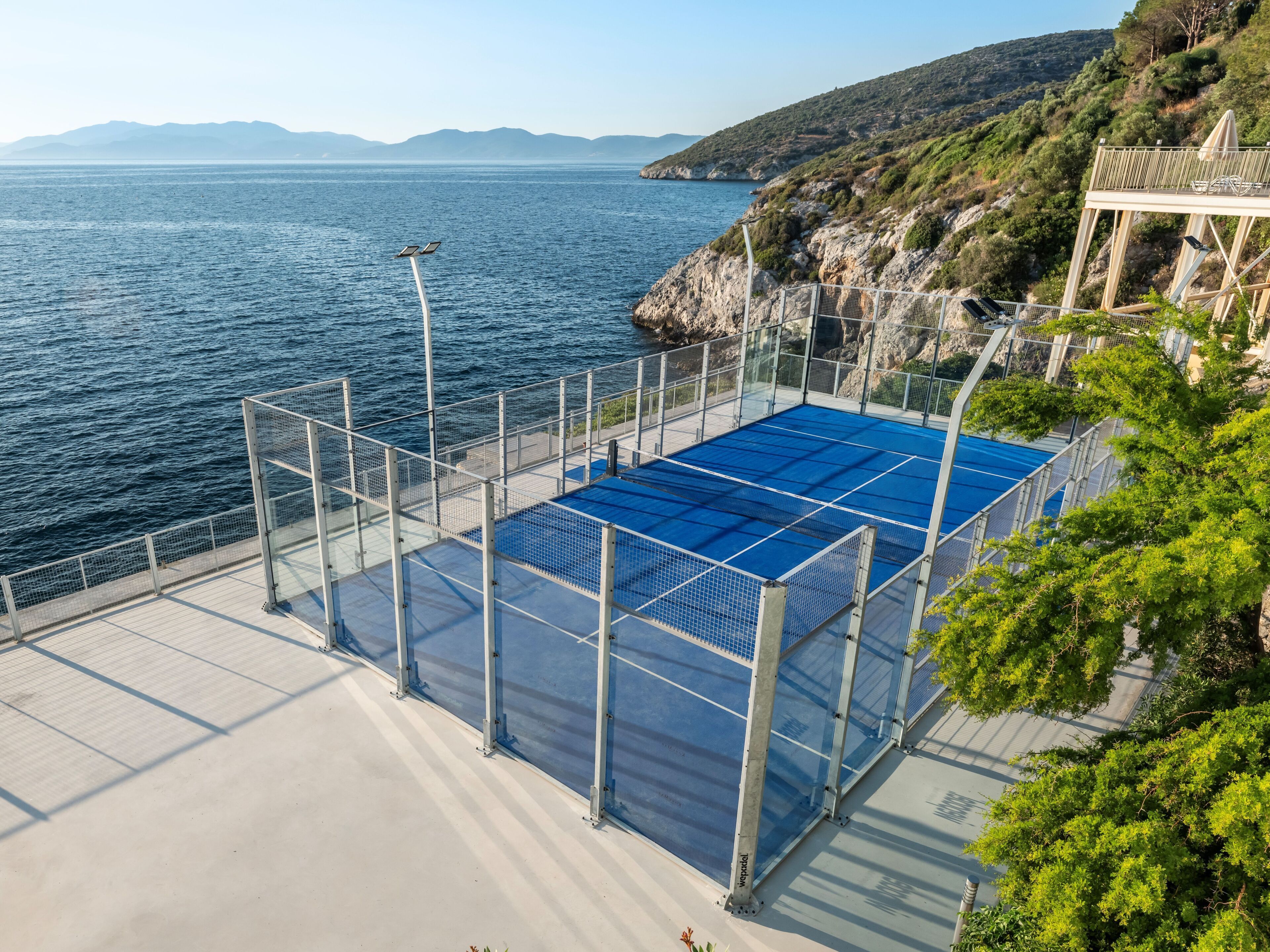 A blue pool with a metal fence, a white deck, and a view of the sea and hills.