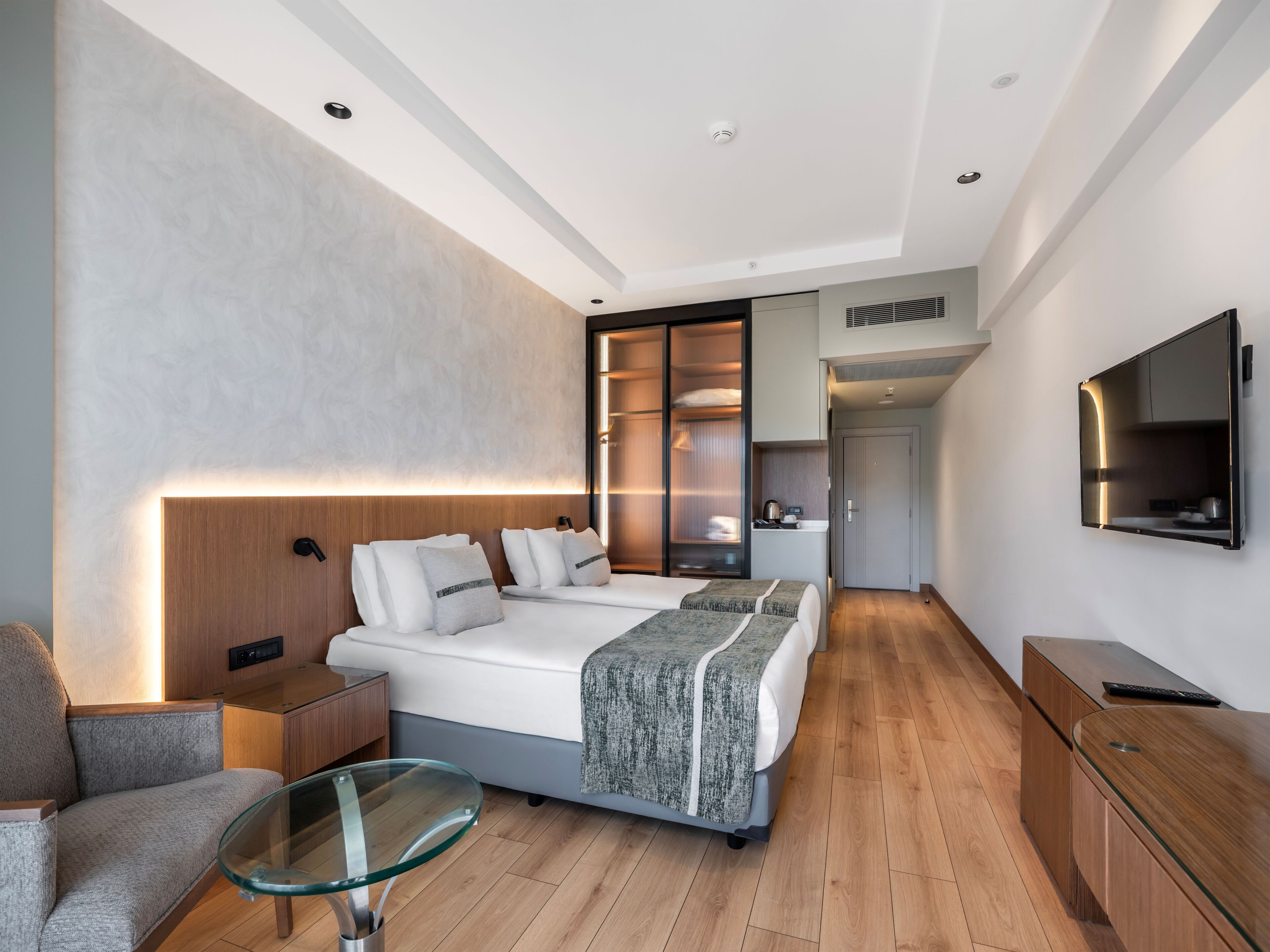A modern hotel room with a large bed, a glass table, a sofa, a wardrobe, and a TV.