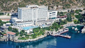 A coastal hotel with a large building, a swimming pool, and a pier extending into the water. - Korumar Deluxe Hotel (Kusadasi)