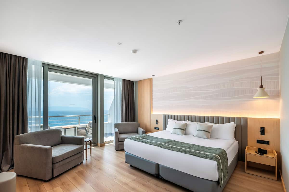 A modern hotel room with a large bed, two armchairs, a desk, and a view of the sea.