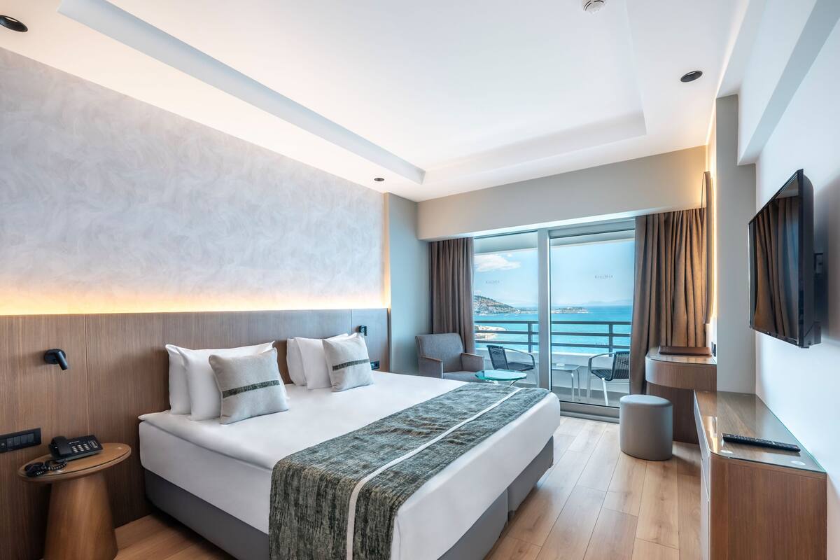 A modern hotel room with a large bed, a desk, a TV, and a balcony with a view.