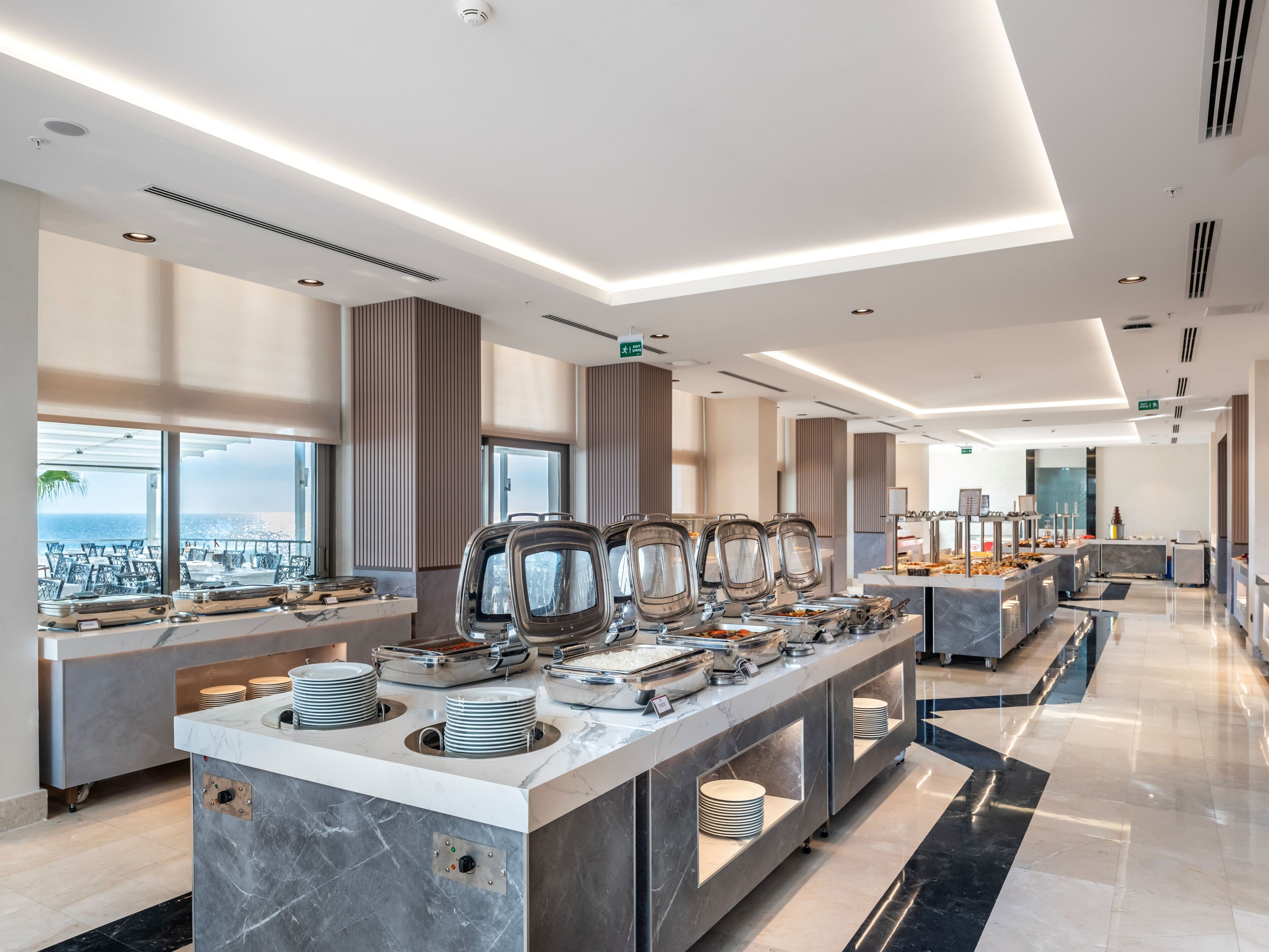 Una moderna presentación de buffet con múltiples estaciones de servicio, una encimera de mármol y vistas a la ciudad a través de grandes ventanales.