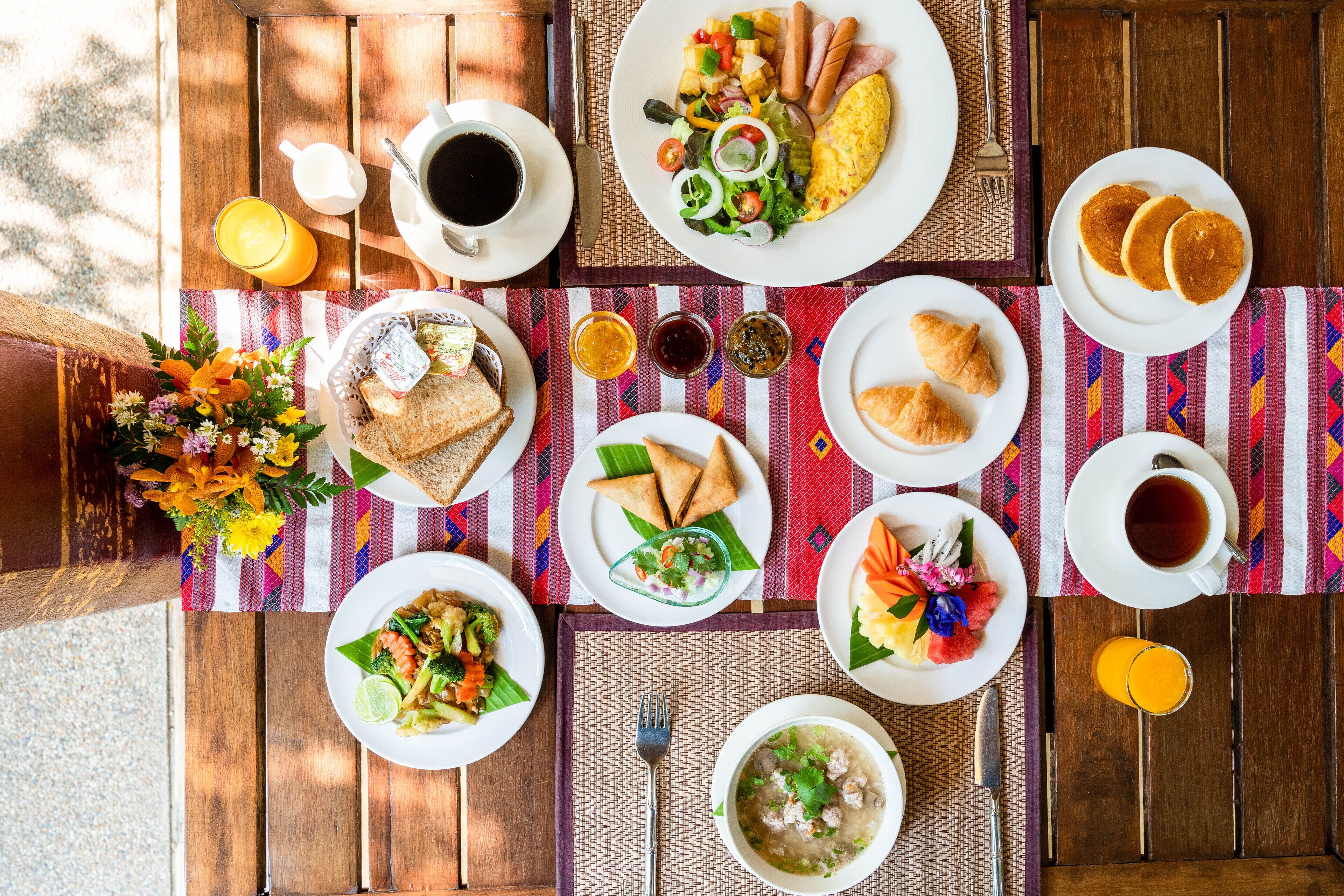 Una mesa de madera con diversos platos para el desayuno, como tortitas, tortillas y ensaladas, junto con tazas de café y zumo.