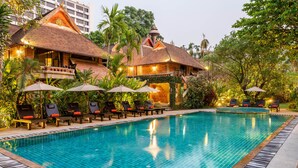 A swimming pool with lounge chairs and umbrellas, surrounded by lush greenery and a traditional building with a tiled roof.