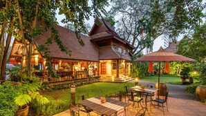 A traditional wooden house with a patio area featuring tables and chairs, surrounded by lush greenery and a well-maintained lawn.