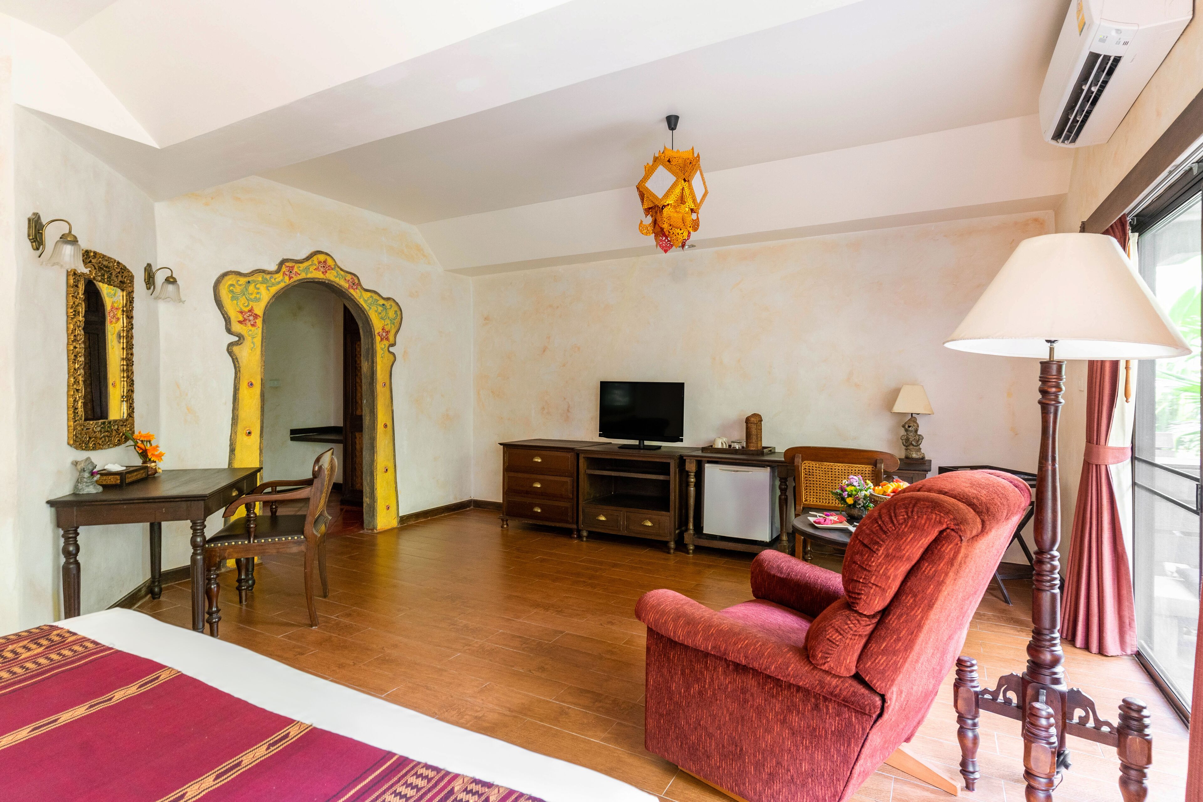 Una habitación con piso de madera, un sillón rojo, un televisor, un mueble pequeño y un espejo decorativo.