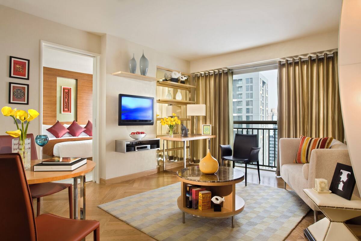 A modern hotel room with a bed, a TV, a dining area, and a balcony with curtains.