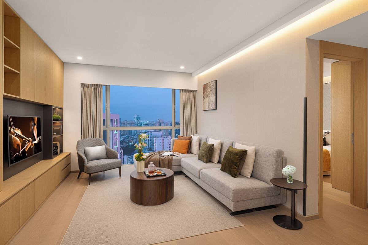 A modern living room with a large TV, comfortable seating, a coffee table, and a view of the cityscape through large windows.