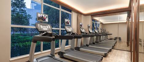 A gym with treadmills aligned against a wall with large windows, offering views of trees and buildings outside.