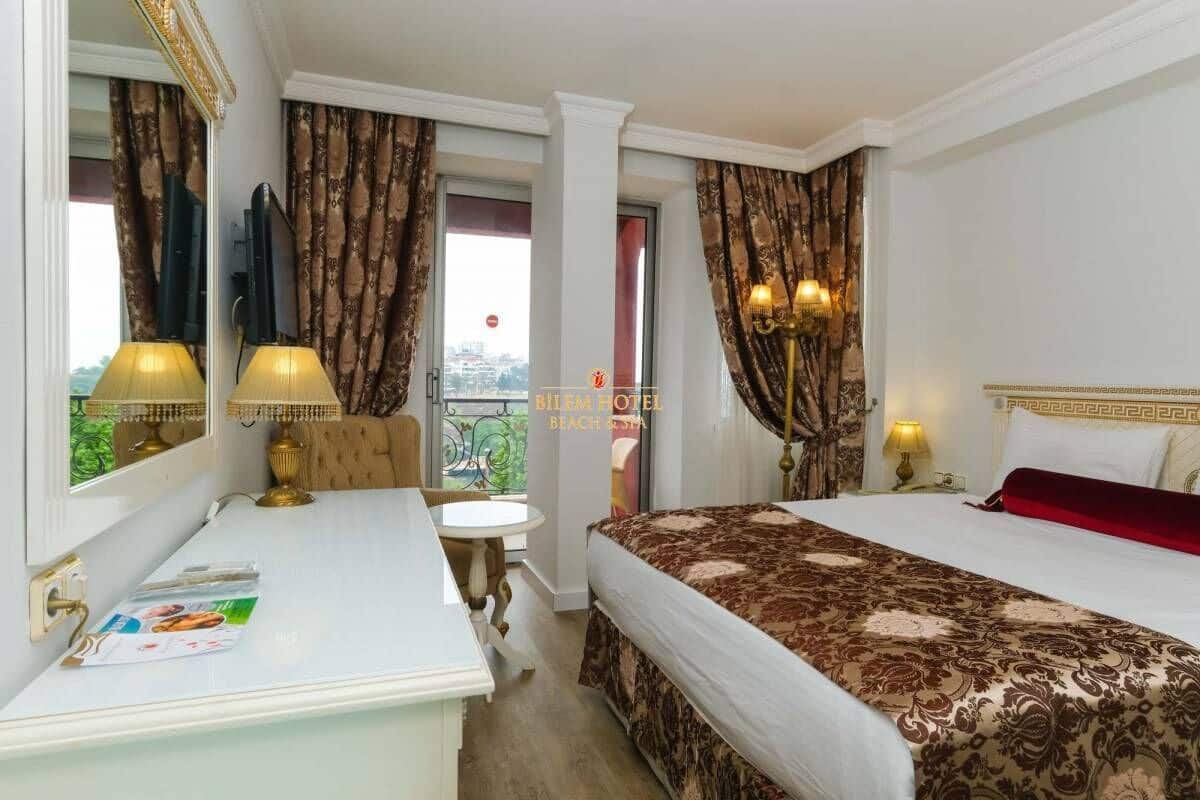 Habitación de hotel con cama, escritorio, silla, lámpara y ventana con cortinas.