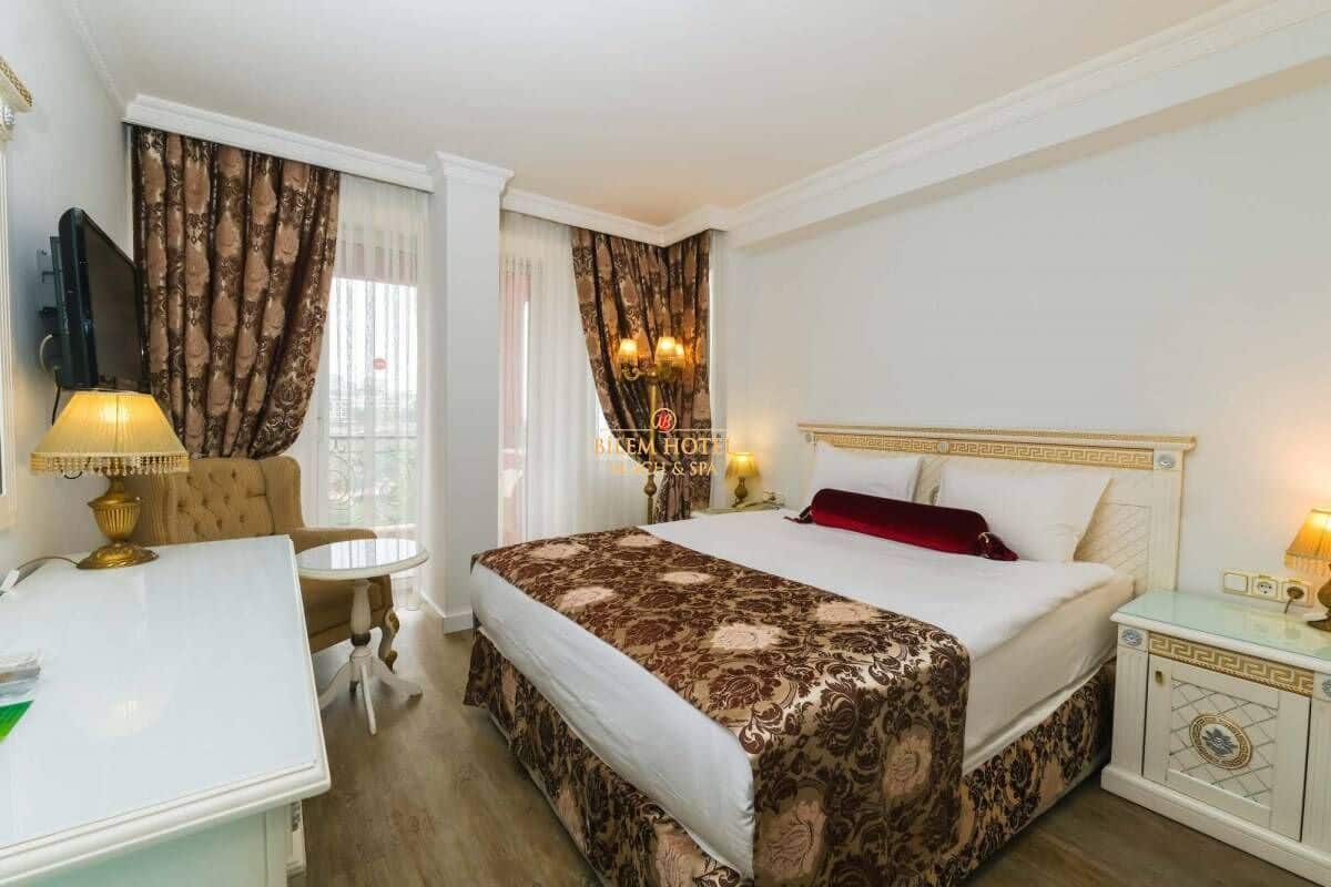 Habitación de hotel con cama, escritorio, silla, televisor y ventana con cortinas.