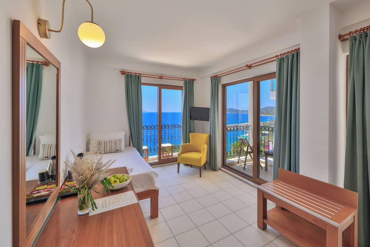 A hotel room with a bed, a desk, a chair, a bench, a TV, and a balcony with a view of the sea.