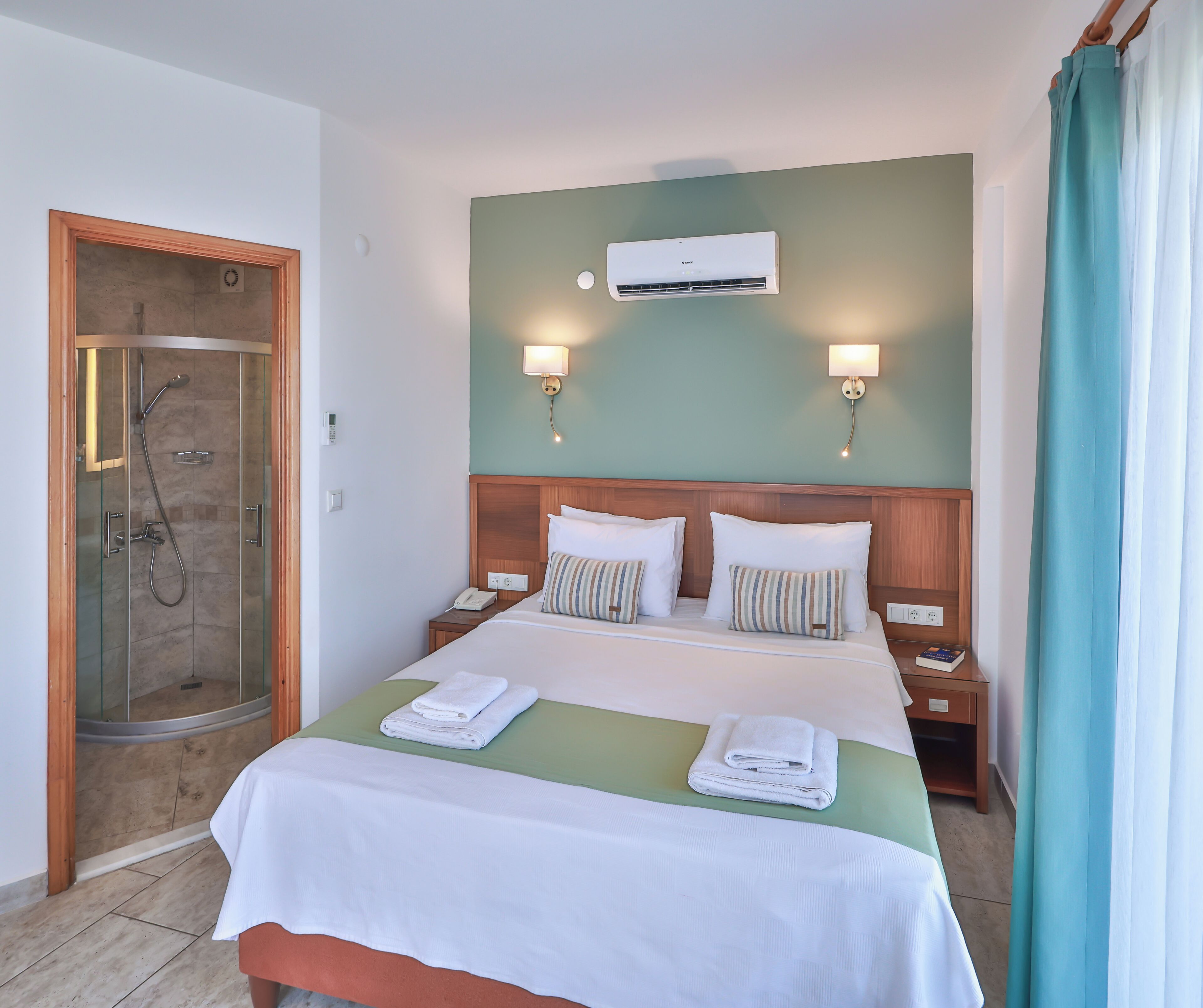 Ein Hotelzimmer mit einem Bett, zwei Nachttischen, einer Klimaanlage, einer Duschkabine und einem Vorhang.
