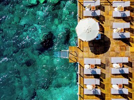 Vista aérea de una terraza de madera con sillones de descanso y sombrillas, con vistas a aguas turquesas cristalinas.