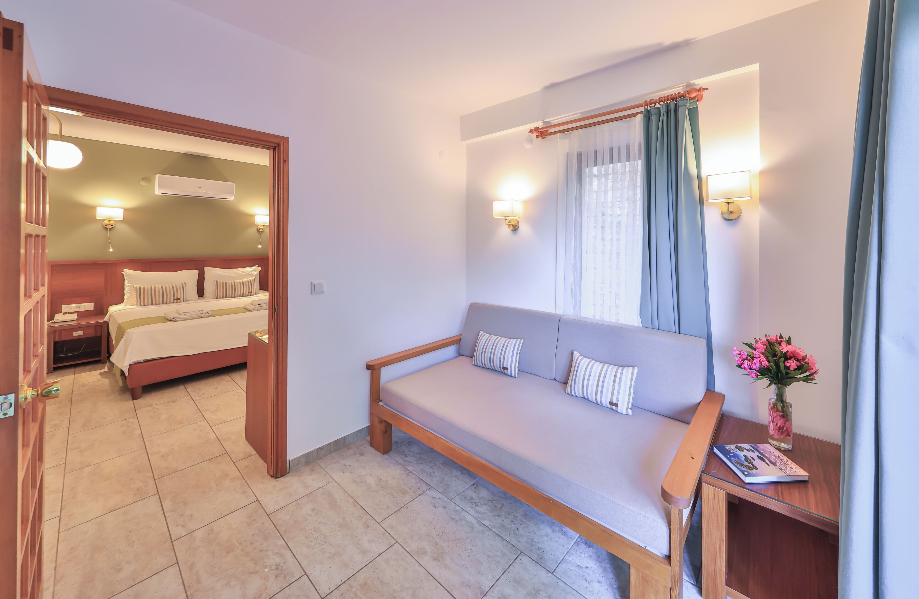 A hotel room with a bed, a sofa, a nightstand, a lamp, and a window with curtains.