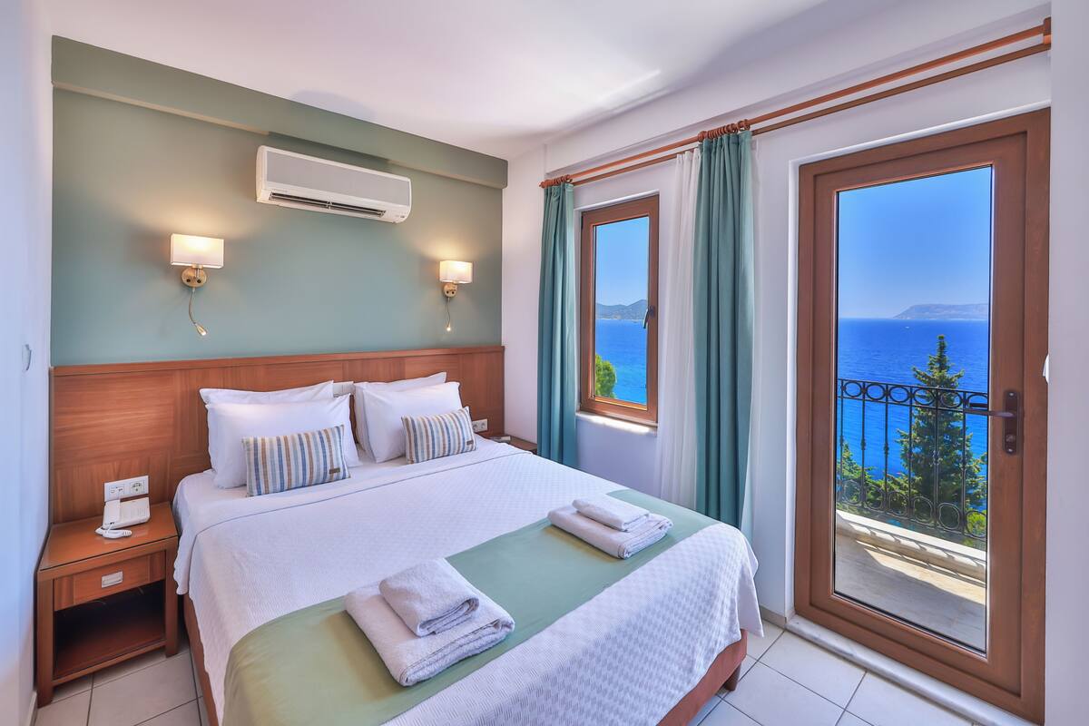A hotel room with a bed, bedside lamps, an air conditioner, a view of the sea, and a balcony with a railing.