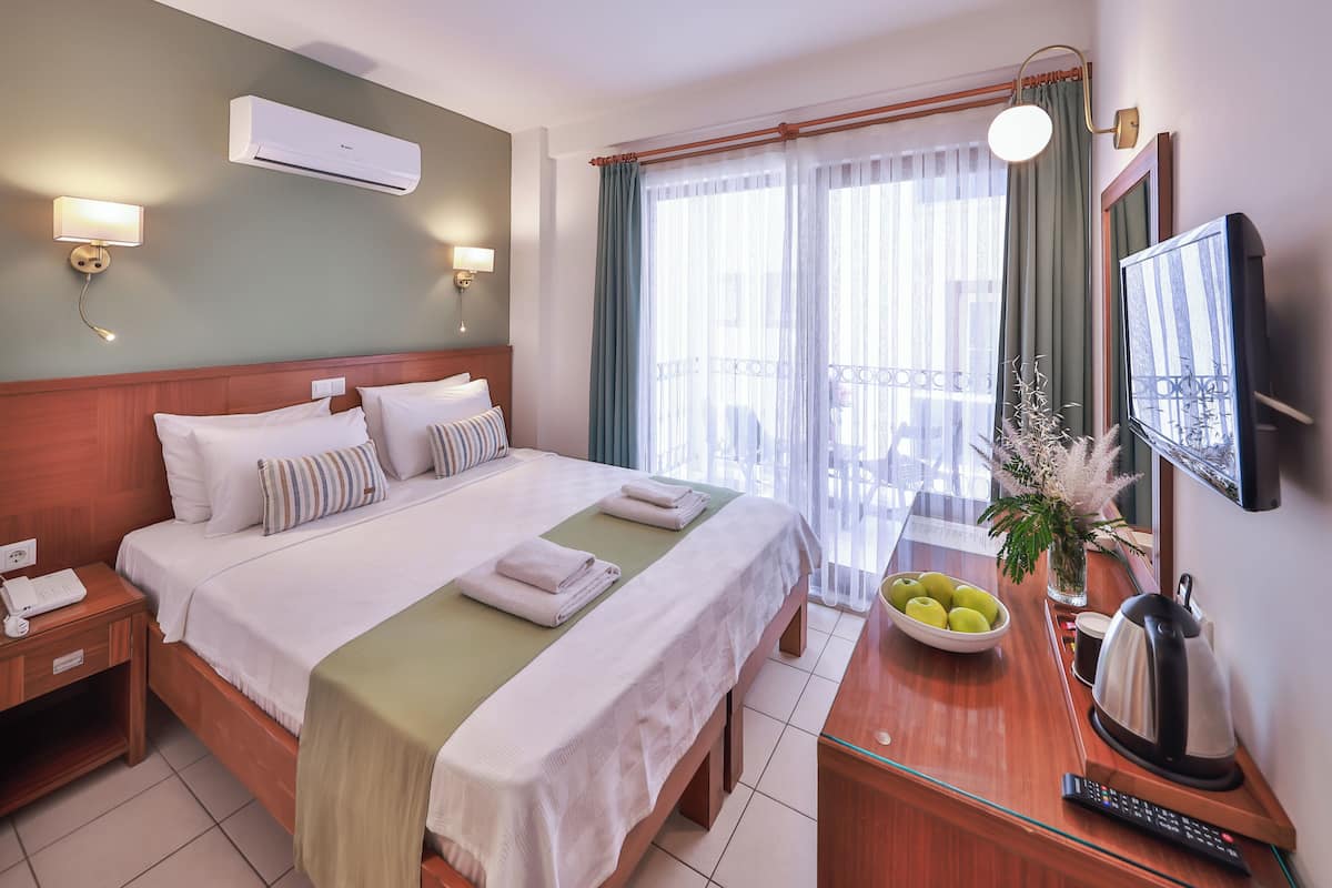 A hotel room with a bed, bedside table, lamp, air conditioning unit, window with curtains, and a television.