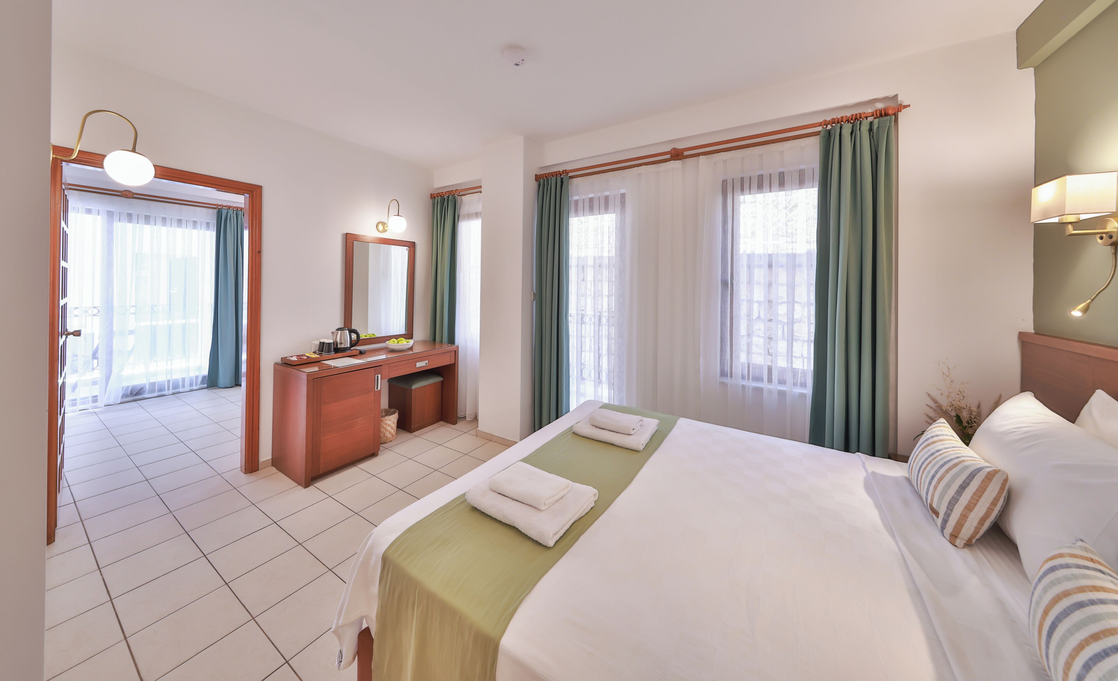 Una habitación de hotel con cama, escritorio, espejo, ventana con cortinas y lámpara.