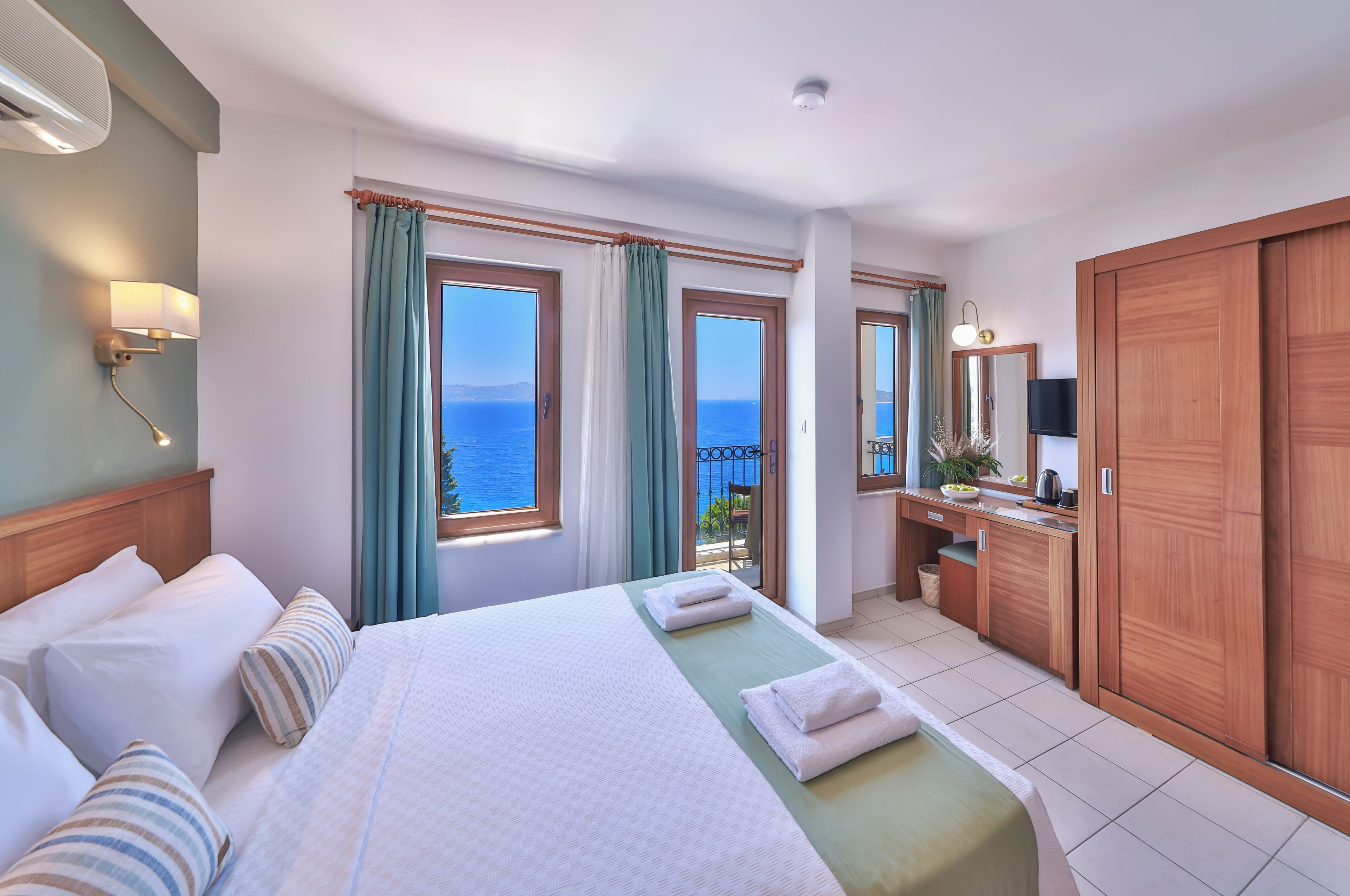 A hotel room with a bed, a desk, a TV, and a view of the sea.