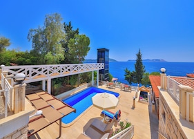 A pool area with lounge chairs, a pergola, and a view of the sea.