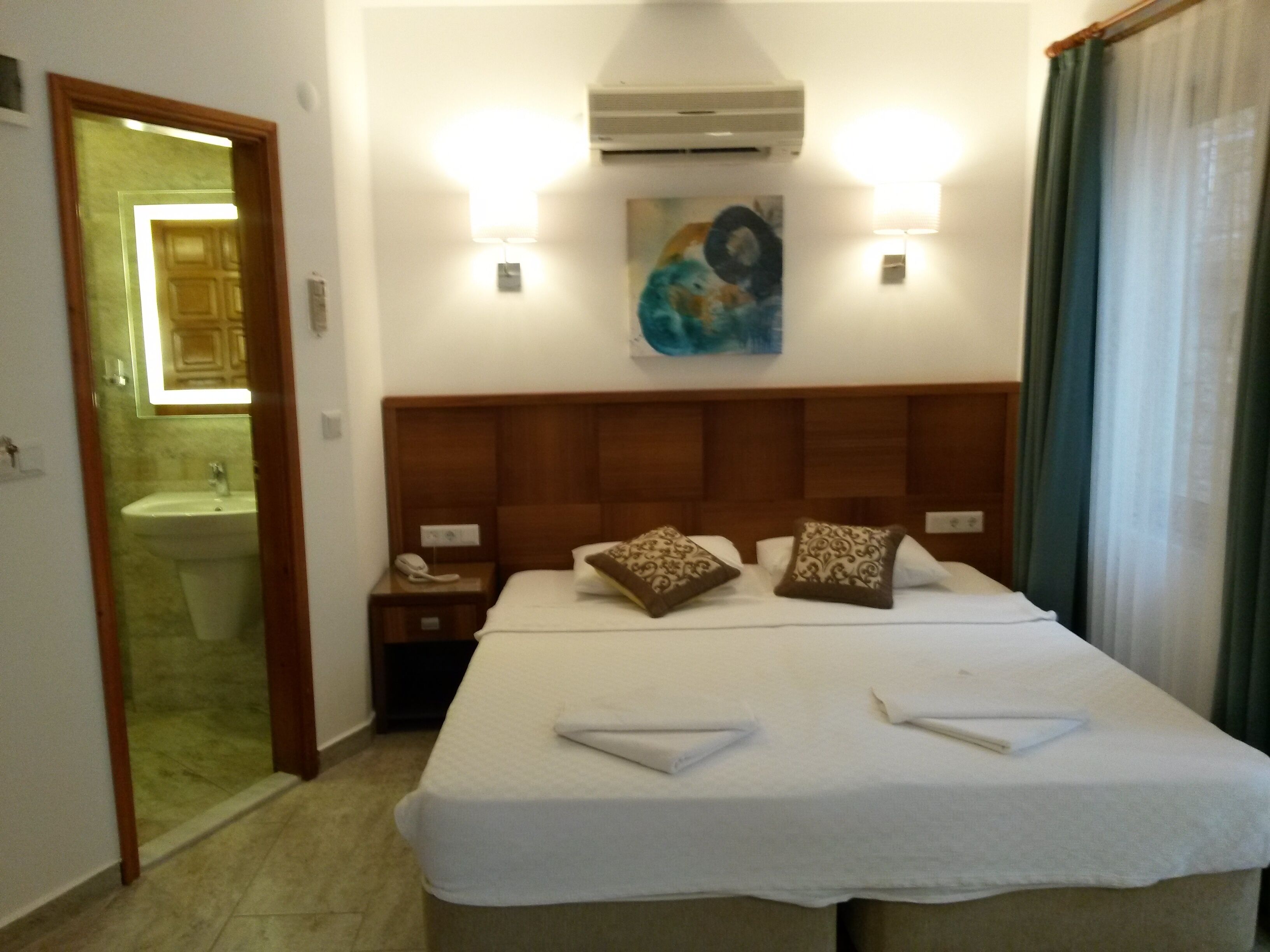 A hotel room with a bed, a nightstand, a painting, an air conditioning unit, and a bathroom visible through an open door.