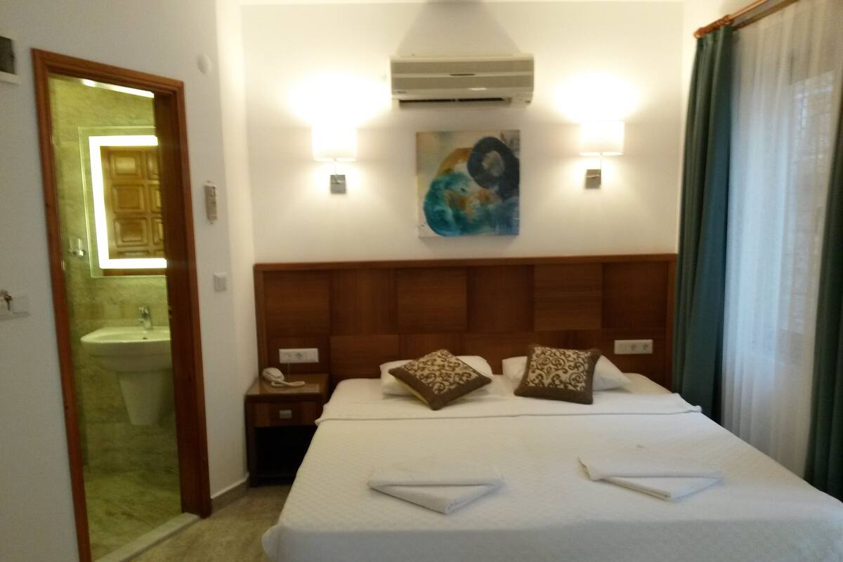 A hotel room with a bed, a nightstand, a painting, an air conditioning unit, and a bathroom visible through an open door.