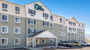 Un hotel de varios pisos con entrada central, unidades de aire acondicionado y un estacionamiento con varios automóviles.