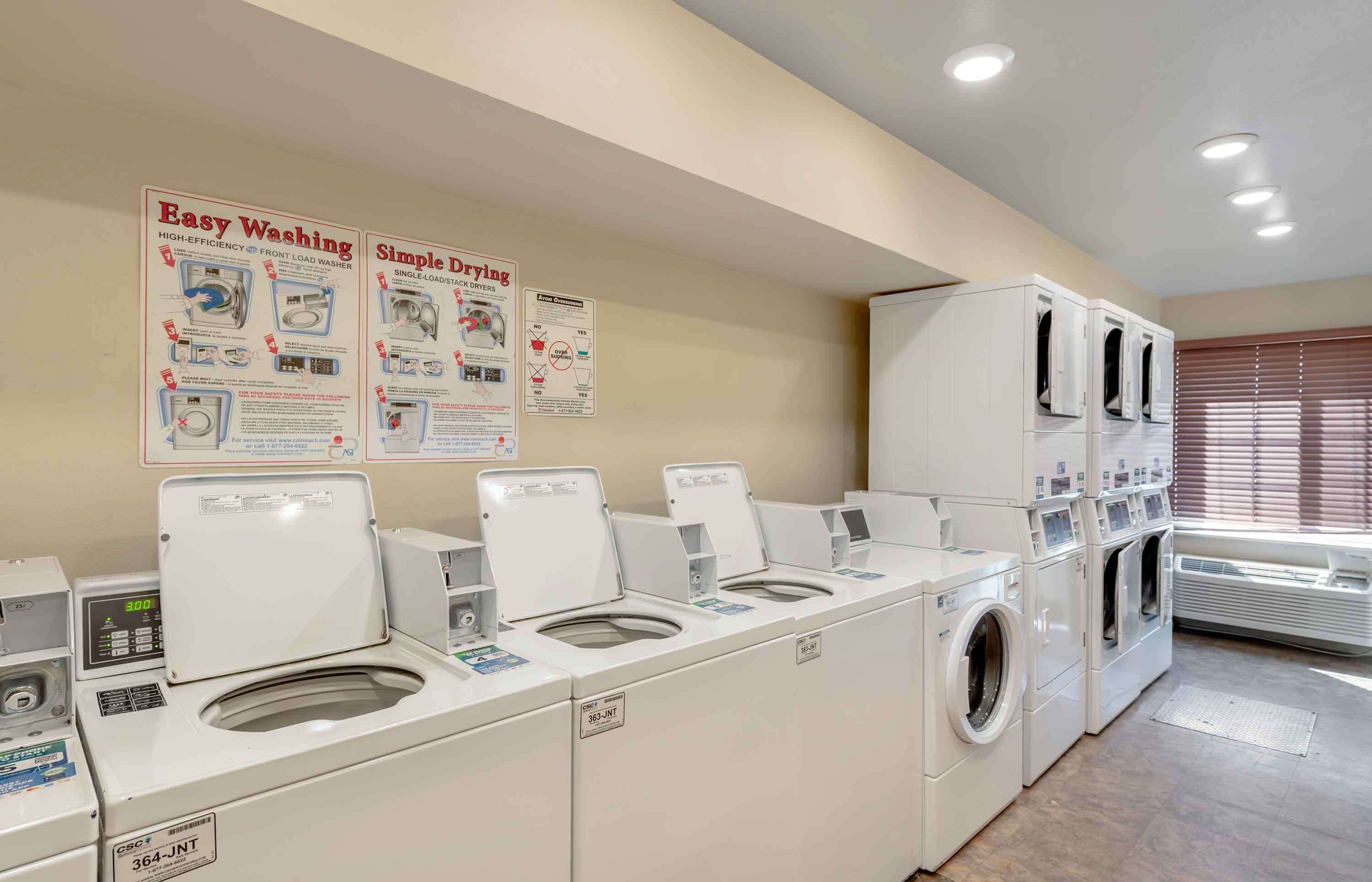 Une buanderie équipée de plusieurs machines à laver et de plusieurs séchoirs, avec des affiches explicatives accrochées au mur et une fenêtre munie de stores.