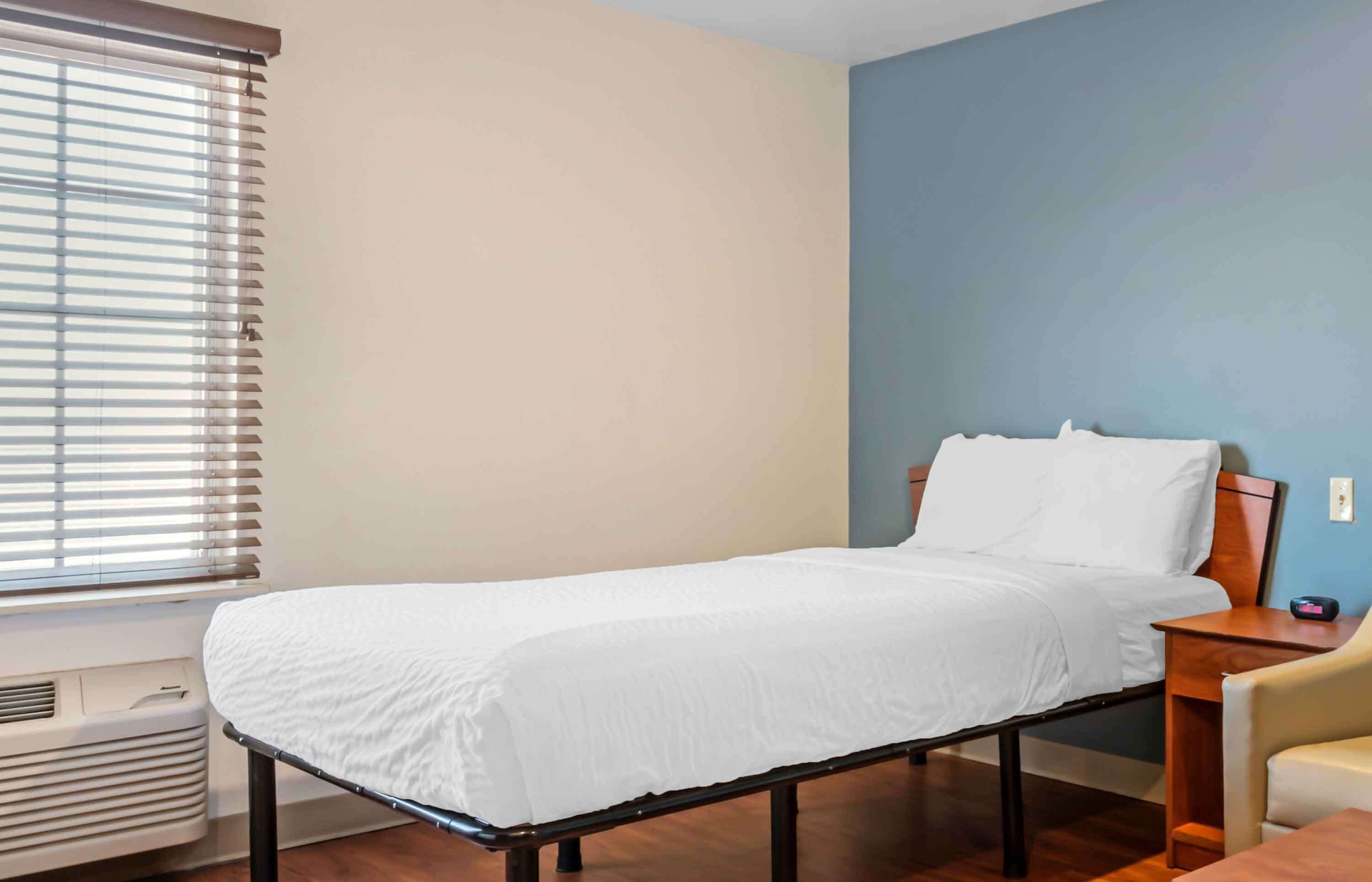 Uma cama de solteiro com colchão e travesseiros brancos, uma mesinha de cabeceira de madeira, uma cadeira e uma janela com persianas.