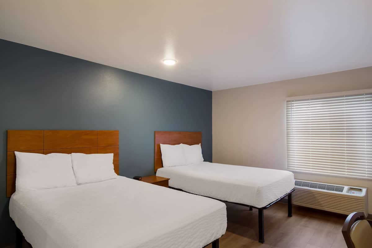 Una habitación de hotel con dos camas individuales, cabecera de madera, una mesita de noche y una ventana con persianas.