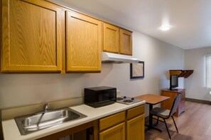 A compact kitchen with wooden cabinets, a stainless steel sink, a microwave, and a small dining table with a chair.