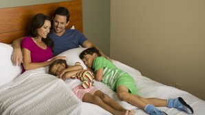 Eine Familie mit drei Personen sitzt auf einem Bett mit einem Holzkopfende, einer weißen, strukturierten Bettdecke und einer schlichten Wand im Hintergrund.