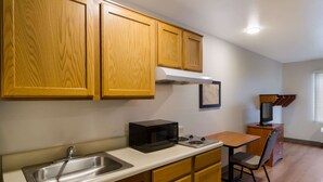 A compact kitchen with wooden cabinets, a microwave, a sink, a small table, and a chair. - WoodSpring Suites Jacksonville East 295 Cruise Port (Jacksonville)