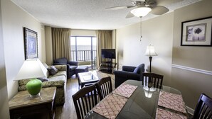A hotel room with a dining table, chairs, a sofa, a television, and a ceiling fan.