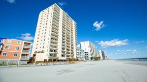A beachfront hotel with multiple floors, balconies, and a sandy beach in the background.