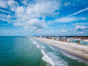 Une plage avec une jetée qui s’avance dans l’océan, bordée de bâtiments et sous un ciel dégagé.