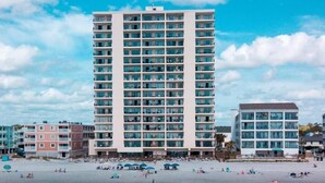 Un hotel frente a la playa con varios pisos, una playa arenosa en primer plano y un cielo despejado.