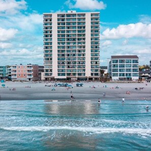Un hotel frente a la playa con varios pisos, una playa arenosa en primer plano y un cielo despejado.
