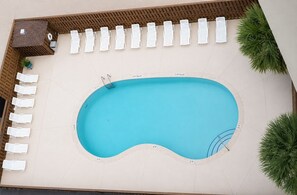 A swimming pool with a wooden deck, surrounded by white lounge chairs, and a small structure with a roof.